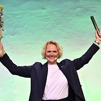 Person i kostym sträcker armarna uppåt med blombukett i ena handen och rulle i andra.