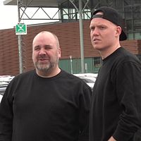 Två personer, män, iklädda svarta tröjor står på en parkeringsplats.