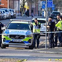 Poliser vid avspärrning med polisbil på stadsgata, flera personer i bakgrunden.