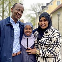 Tre personer står tillsammans utomhus, med stenvägg och byggnad i bakgrunden.