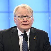 Peter Hultqvist talar i en mikrofon framför en blå bakgrund med Sveriges riksdags logotyp.