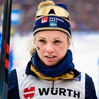 Skidåkare i tävlingsdräkt håller stav på snötäckt bana.