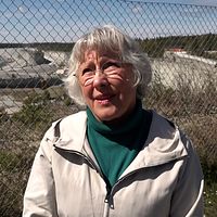 Person med vitt hår står utomhus vid stängsel, hav och industriområde syns i bakgrunden.