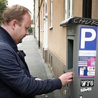 En person står vid en parkeringsautomat och använder den, med ett parkeringstecken synligt på automaten. I bakgrunden syns en rad med parkerade bilar och en gata kantad av träd.