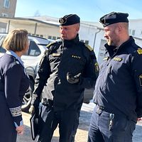 Uniformerad personal diskuterar utomhus framför byggnader. En bil syns i bakgrunden.