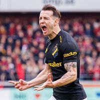 Fotbollsspelare i svart tröja firar framför publik på läktaren.