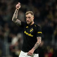 Fotbollsspelare i AIK-tröja höjer handen under match, suddiga åskådare i bakgrunden.