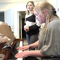 Två tjejer vid ett piano. En spelar och en sjunger.