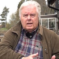 En person i rutskjorta sitter utomhus vid ett trästaket, med en röd fågelholk i bakgrunden.