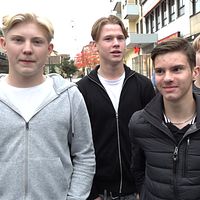 En grupp unga män står på en gata, omgivna av byggnader och affärer. De ser ut att vara mitt i en social interaktion, klädda i vardagliga kläder.