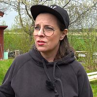 Person med keps utanför ett rött hus med trädgårdsslang och grönska i bakgrunden.