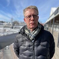 Per Högberg (KD) i vinterjacka står vid snötäckt park, med en solig himmel och promenadstråk i bakgrunden.