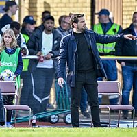 Två fotbollstränare ger instruktioner vid sidlinjen under en match, publik i bakgrunden.