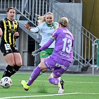 Fotbollsmålvakt i lila räddar skott mot två backar i svartgula tröjor och en anfallare.