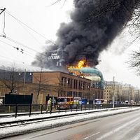 Delad bild. Vänster: person talar vid podiet. Höger: byggnad i brand med rök.