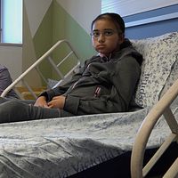 Amira ligger i sjukhussäng, hennes mamma sitter bredvid sängen.