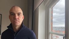 Stefan Dalteg står vid ett fönster med utsikt över stadsmiljö. Han tittar allvarligt in i kameran.