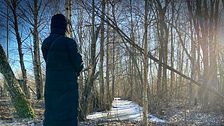 Person i lång kappa står vid snötäckt stig i skog med solljus i bakgrunden.