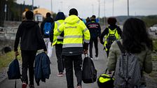 Flera Northvoltarbetare med väskor promenerar längs en väg, några bär reflexjackor där det står Northvolt på ryggen.