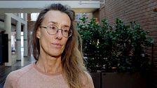 En person står inomhus med en växtfylld bakgrund av tegelväggar och en korridor. Personen bär en ljusrosa tröja med geometriskt mönster.