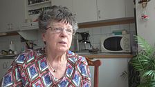 En äldre kvinna sitter i ett hemtrevligt kök med mikro och växter i bakgrunden.