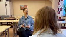 Två personer sitter vid skolbänkar och har ett samtal i ett klassrum.