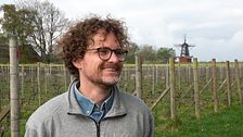 En man står framför en vingård på Kullaberg. I bakgrunden syns en vinodling och en kvarn.