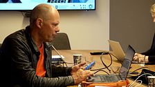 medelålders man med rakat huvud sitter med mobiltelefon framför sin dator i ett konferensrum
