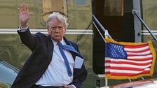 En man i kostym vinkar bredvid ett amerikanskt flagg framför ett helikopterplan.