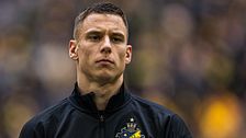 AIK-spelare med mörkt jacka och klubbmärke synligt, suddig publik i bakgrunden.
