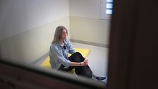 Kvinna sitter ensam på madrass i cell med kala väggar och fönster med galler.