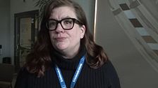 Person med långt brunt hår bär svart tröja och blå namnskylt i en inomhusmiljö.