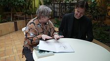 Två personer vid ett bord, diskuterar en bok med illustrationer.