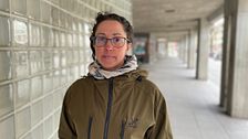 Person i grön jacka står i glasväggskorridor på trottoaren.
