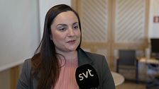 Kvinna i rosa tröja med SVT-mikrofon, stol och bord i bakgrunden.