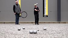 Två personer spelar boule inomhus mot en grå vägg med reklam.