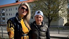 Två personer står utomhus, en kvinna i gul klänning och ett barn i keps. Bakgrund med byggnad.