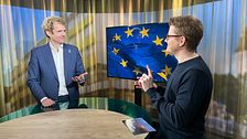 Alexandre Bloxs och Magnus Bergevin står vid ett bord i en studio. Tv-skärmen bakom visar EU-flaggan.
