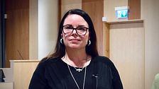 Stina Michelson (S) står i en kommunsal, mikrofon och bänkar syns i bakgrunden.