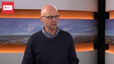 En person står i en tv-studio med panoramabild som bakgrund.