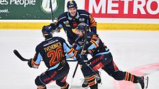 Tre hockeyspelare jublar och kramas på isen efter mål. Skridskor, klubbor och matchdräkter syns.