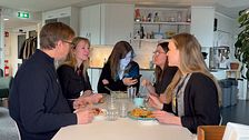Fem personer samtalar runt ett bord i ett kök. En kvinna tittar på sin mobiltelefon.