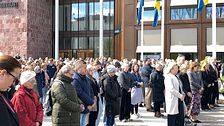 Stor folkmassa står samlad utanför en byggnad med svenska flaggor i solsken.