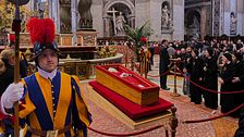 Soldat vaktar vid påvens kista under ceremoni i en utsmyckad kyrka, sörjande i bakgrunden.