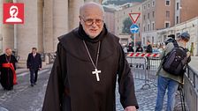 En präst i svart kåpa och korshalsband går utanför en kyrkobyggnad. Andra personer syns i bakgrunden.