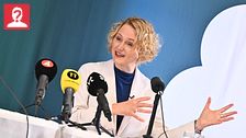En person i kritvit kavaj håller en presskonferens med mikrofoner på bordet.