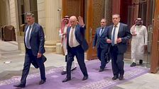 Fyra tjänstemän och två arabiska män går på en lila matta i en lyxig byggnad.