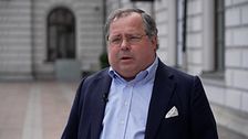 Man i kostym står utomhus framför byggnad med stenväggar och stora fönster.