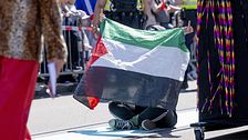 Person sitter på marken och håller upp en palestinsk flagga under en folksamling utomhus.