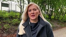 Kvinna med långt blont hår står utomhus på gångväg med gröna träd i bakgrunden.